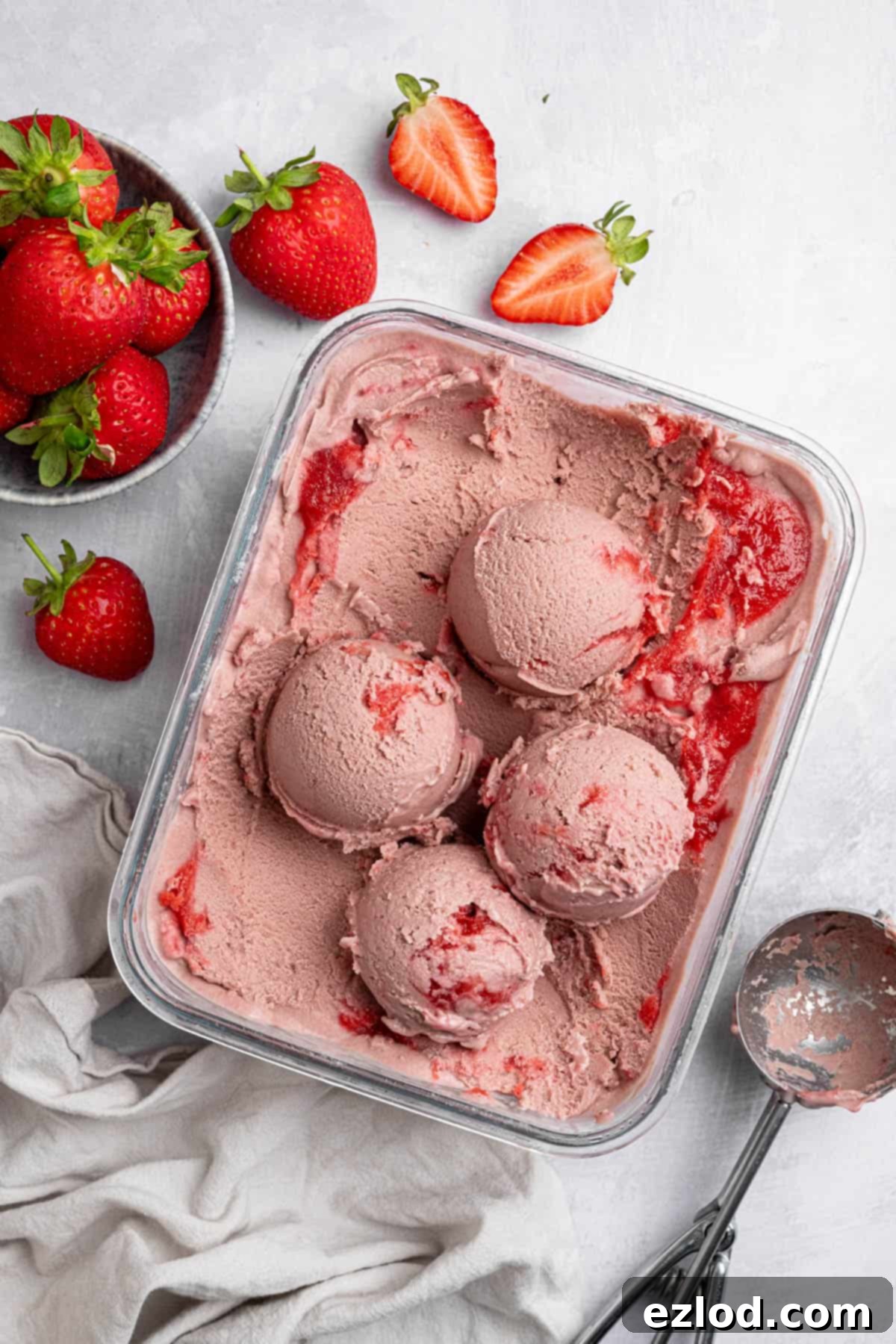 A glass container of vegan strawberry ice cream, some of it scooped into balls, with an ice cream scoop and a bowl of fresh strawberries.