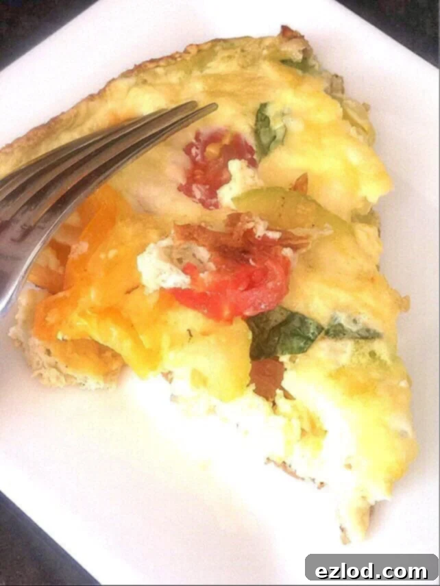 A triangular slice of fritatta on a white plate with tomatoes, zucchini, and basil peeking through the egg dish