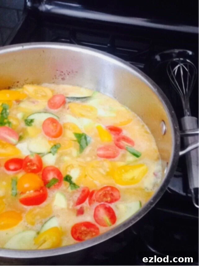 Red and yellow tomatoes, basil, and zucchini begin to cook in a pan with beaten eggs. A whisk lays to the side.