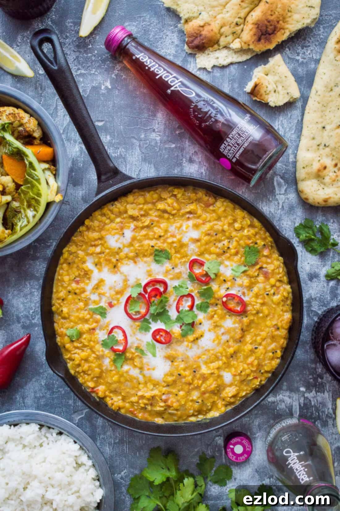 A bubbling pot of vegan red lentil dhal, garnished with fresh red chilli, coriander, and a swirl of coconut milk, with a bottle of Appletiser in the background.