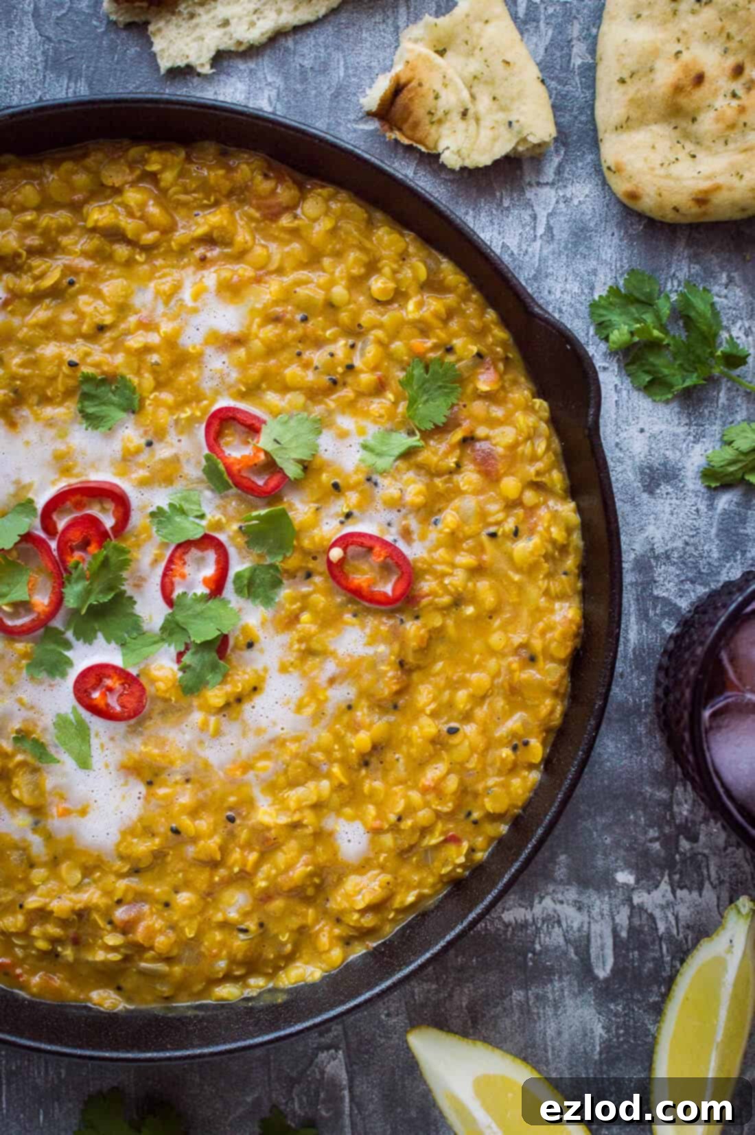 A close-up view of a pan of rich vegan red lentil dhal, generously topped with vibrant red chilli, fresh coriander, and a drizzle of creamy coconut milk.