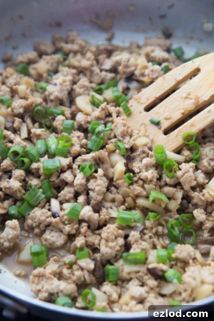 Ground chicken and veggies being sauteed in a pan with a slotted wooden spoon and garnished with green onions