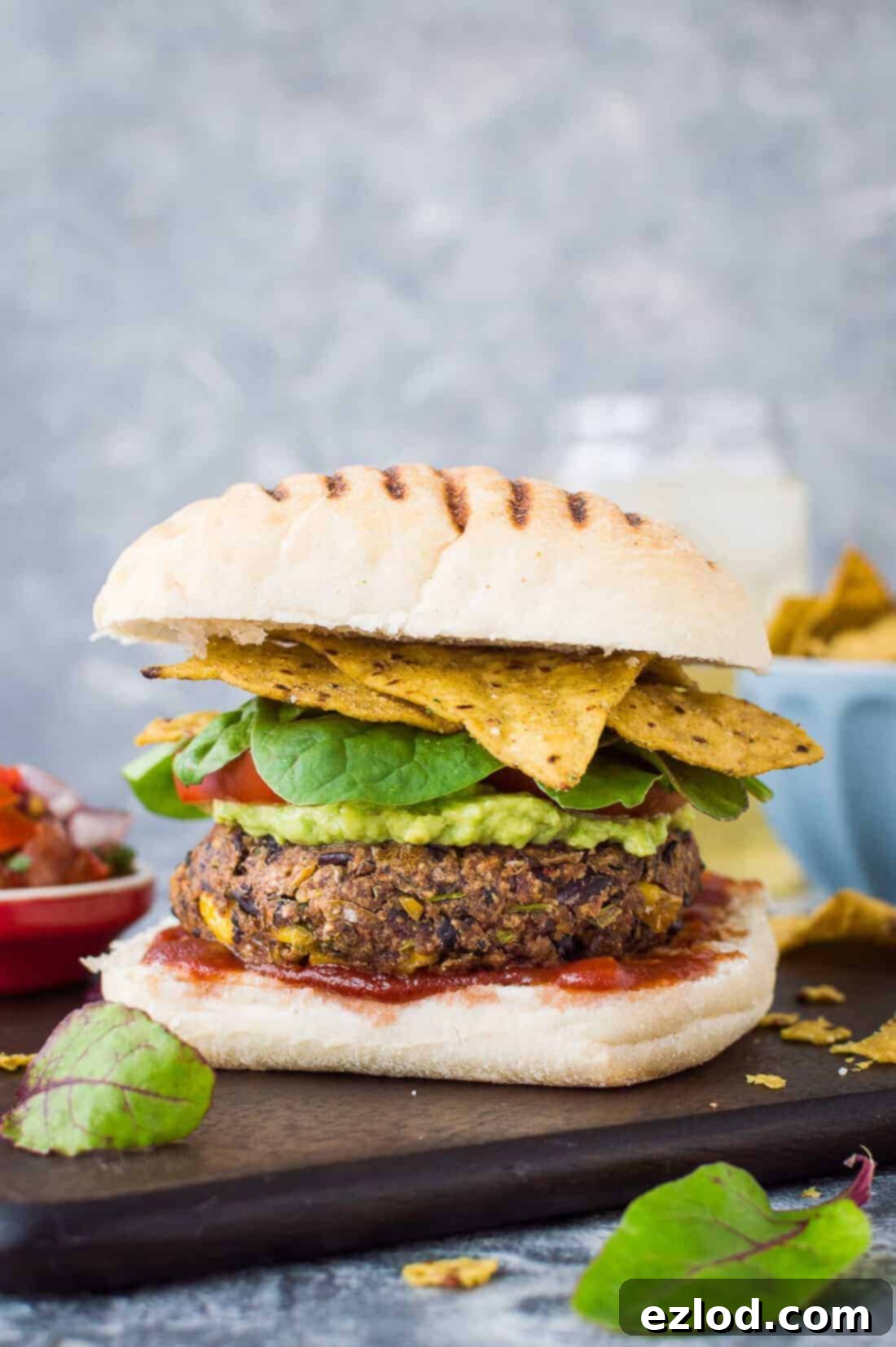 Spicy black and kidney bean burger patty on a bun with guacamole, salsa, and tortilla chips, served alongside an Appletiser bottle. Perfect vegan meal.