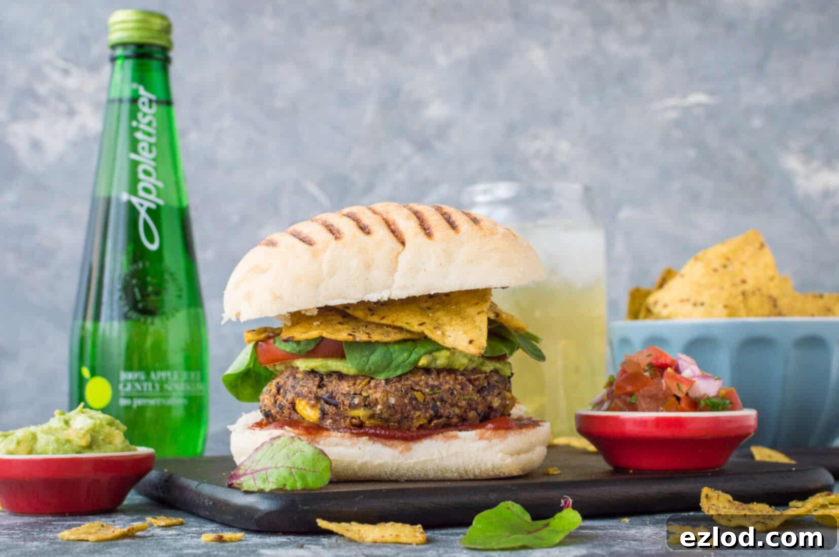 Vegan Mexican bean burger nestled in a toasted bun on a rustic wooden board, accompanied by crunchy tortilla chips, fresh salsa, and an ice-cold Appletiser.