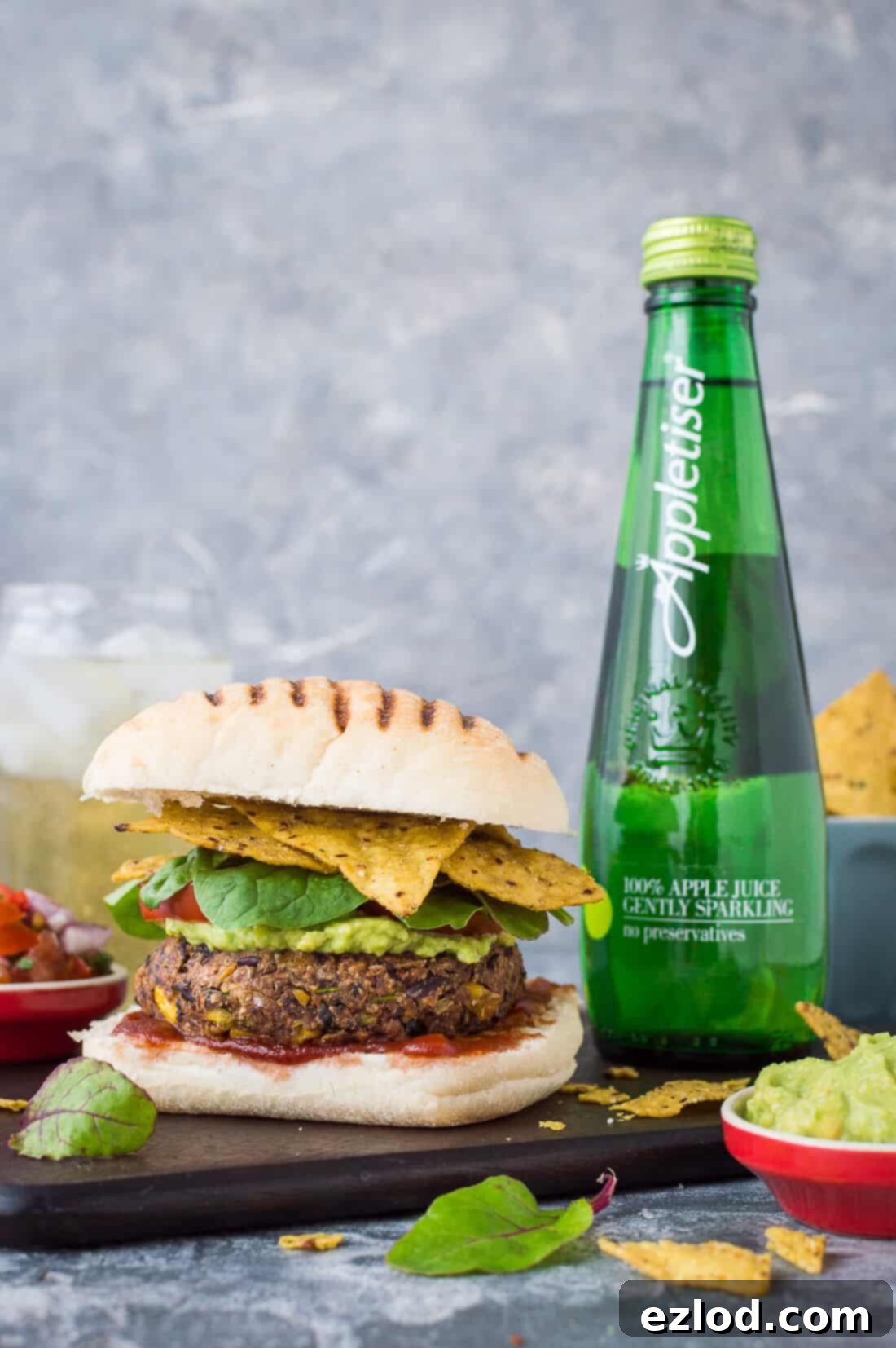 Vegetarian bean burger feast on a wooden board, complete with crispy tortilla chips, vibrant guacamole, zesty salsa, and a refreshing bottle of Appletiser.