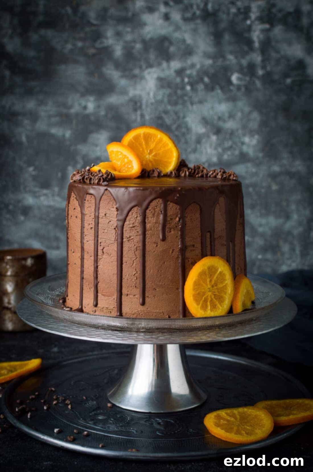 Vegan chocolate, orange and almond cake topped with chocolate drip and candied orange slices on a metal cake stand with a grey background