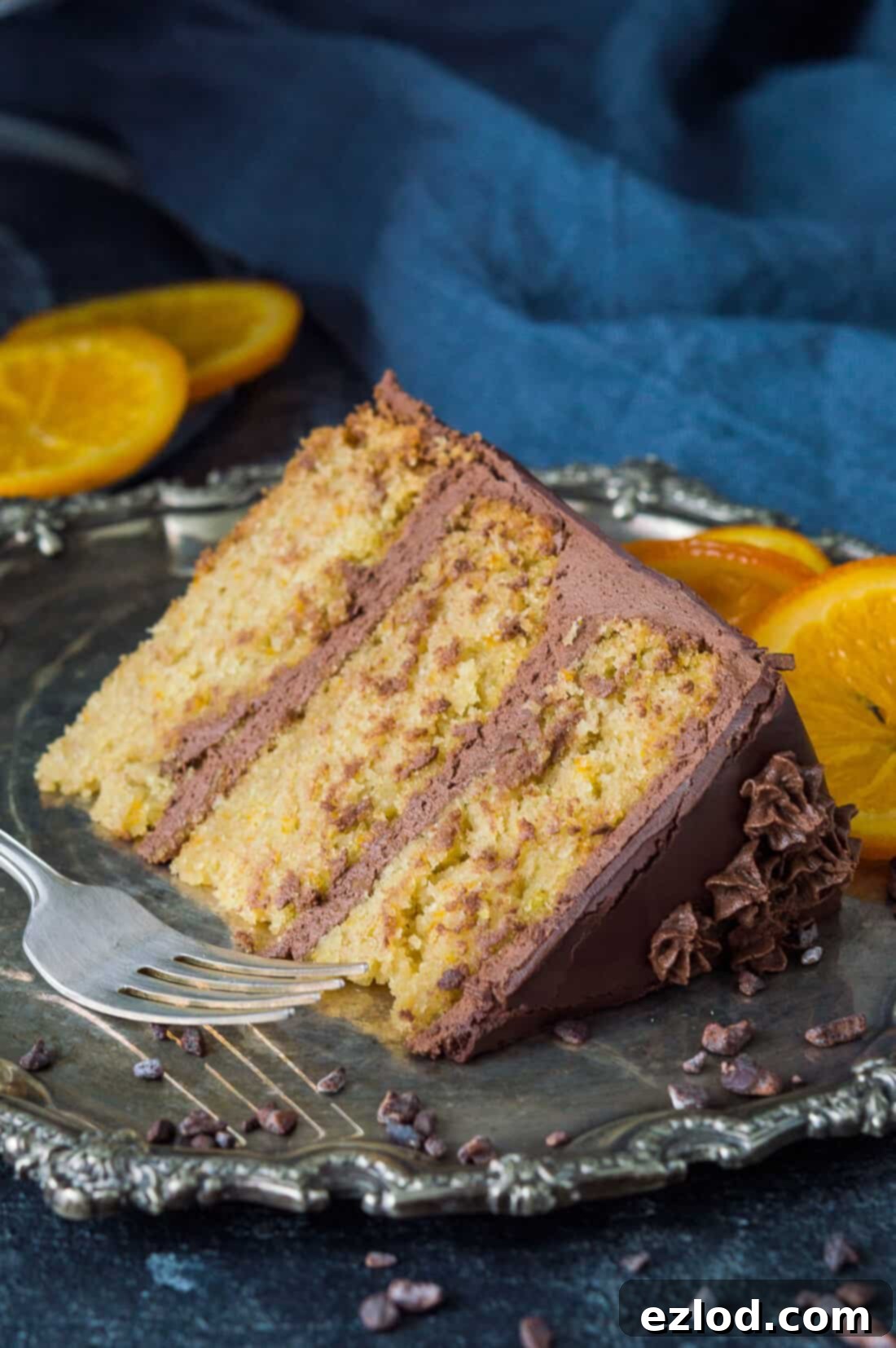 A slice of vegan orange and almond cake with vegan chocolate orange buttercream and candied oranges on a pewter plate.
