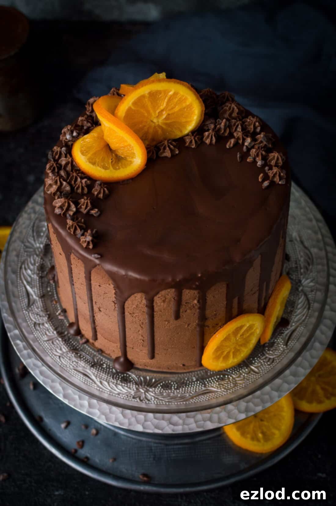 Angled photo of vegan orange and almond cake with chocolate orange vegan buttercream, chocolate drip and candied oranges on a glass plate and silver cake stand.