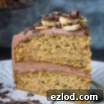 A slice of two-layered vegan banana cake with chocolate peanut butter buttercream, banana chips and chocolate curls on a white plate