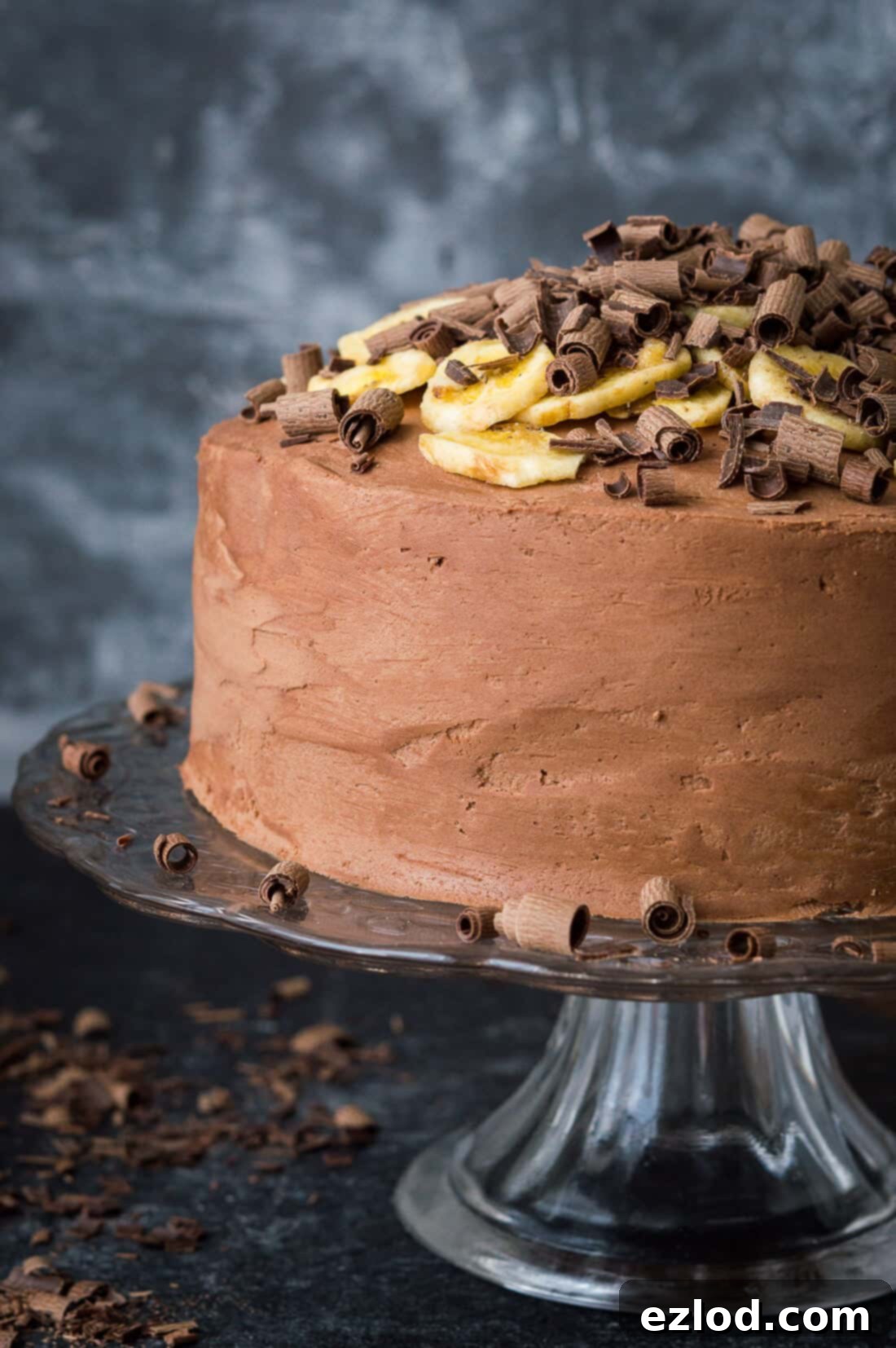 Side shot of vegan banana cake with chocolate peanut butter frosting topped with banana chips and chocolate curls on a glass cake stand.