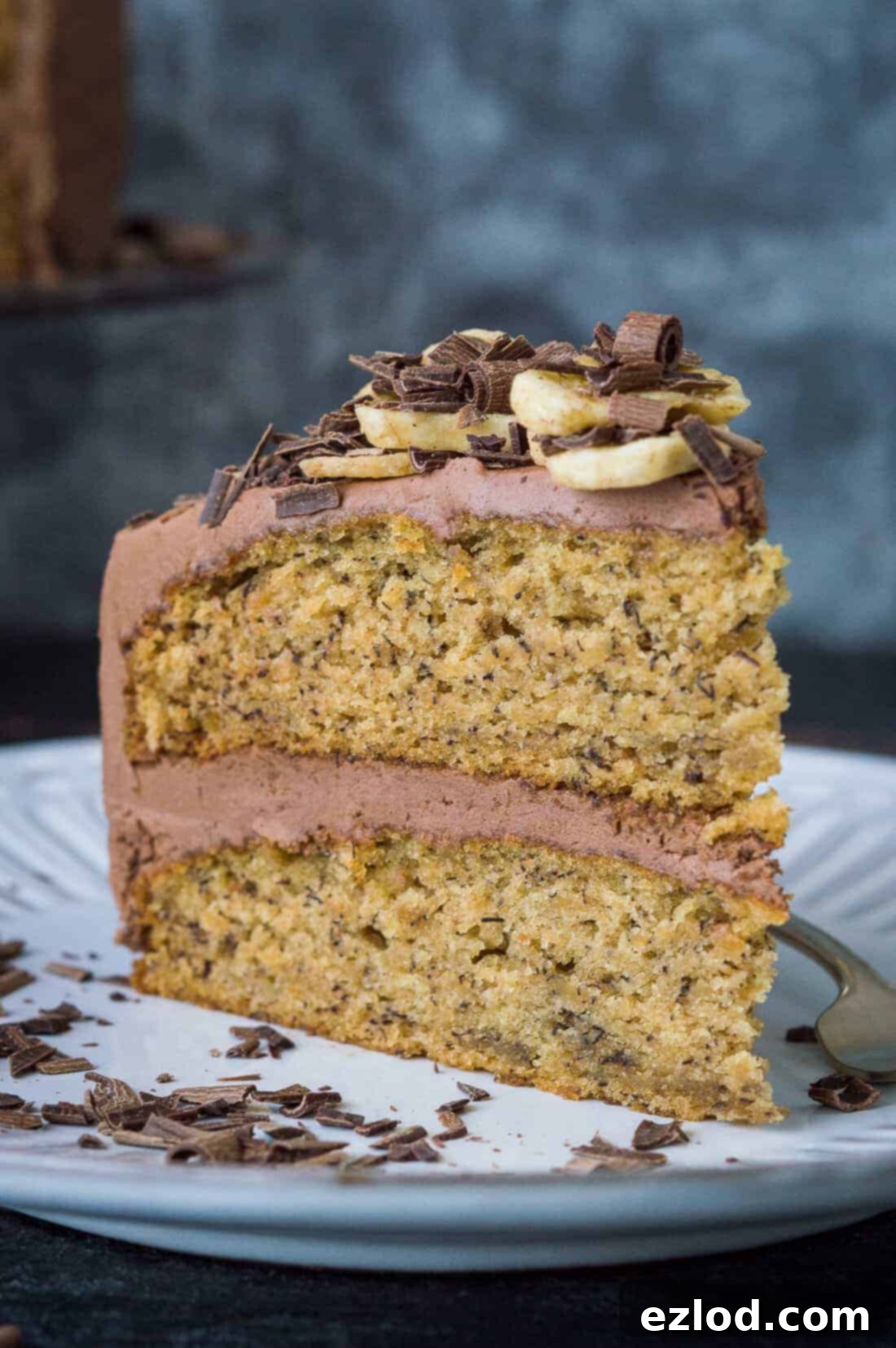 A slice of two-layered vegan banana cake with chocolate peanut butter buttercream, banana chips and chocolate curls on a white plate