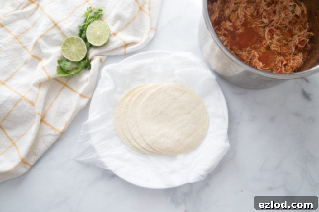 A stack of corn tortillas on a marble surface next to fresh limes and a bowl of shredded salsa lime chicken, showcasing the main components for easy taquito assembly.