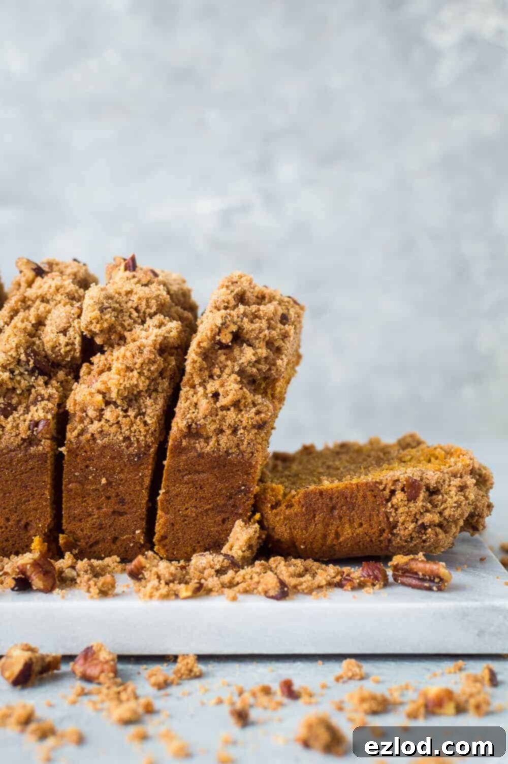 A side view of a perfectly sliced vegan sweet potato loaf cake, revealing its moist interior and generous pecan streusel topping, presented on a marble board with a subtle grey background.