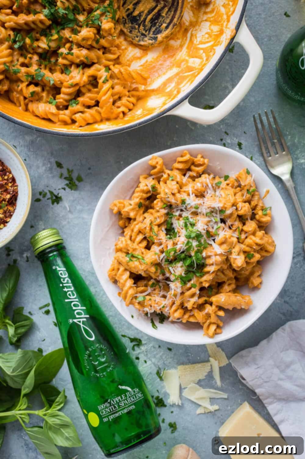 A bowl of vegan creamy roasted red pepper tomato pasta with a bottle of Appletiser.