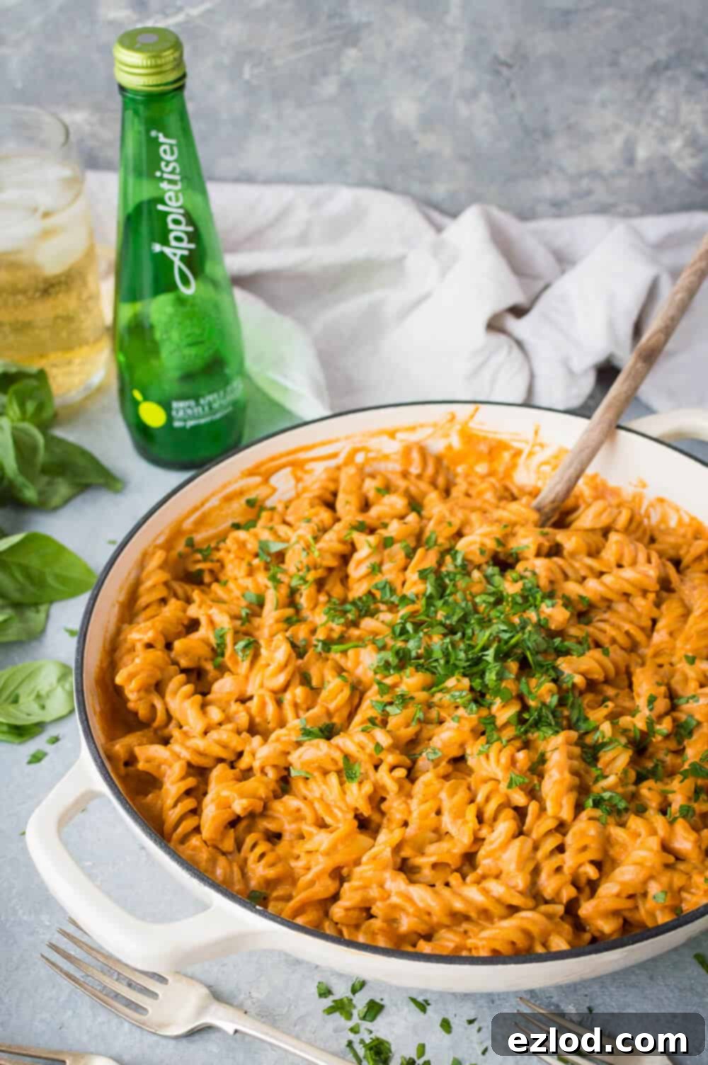 Side angle shot of a pan of vegan creamy roasted red pepper tomato pasta on a grey surface with Appletiser and herbs in the background.