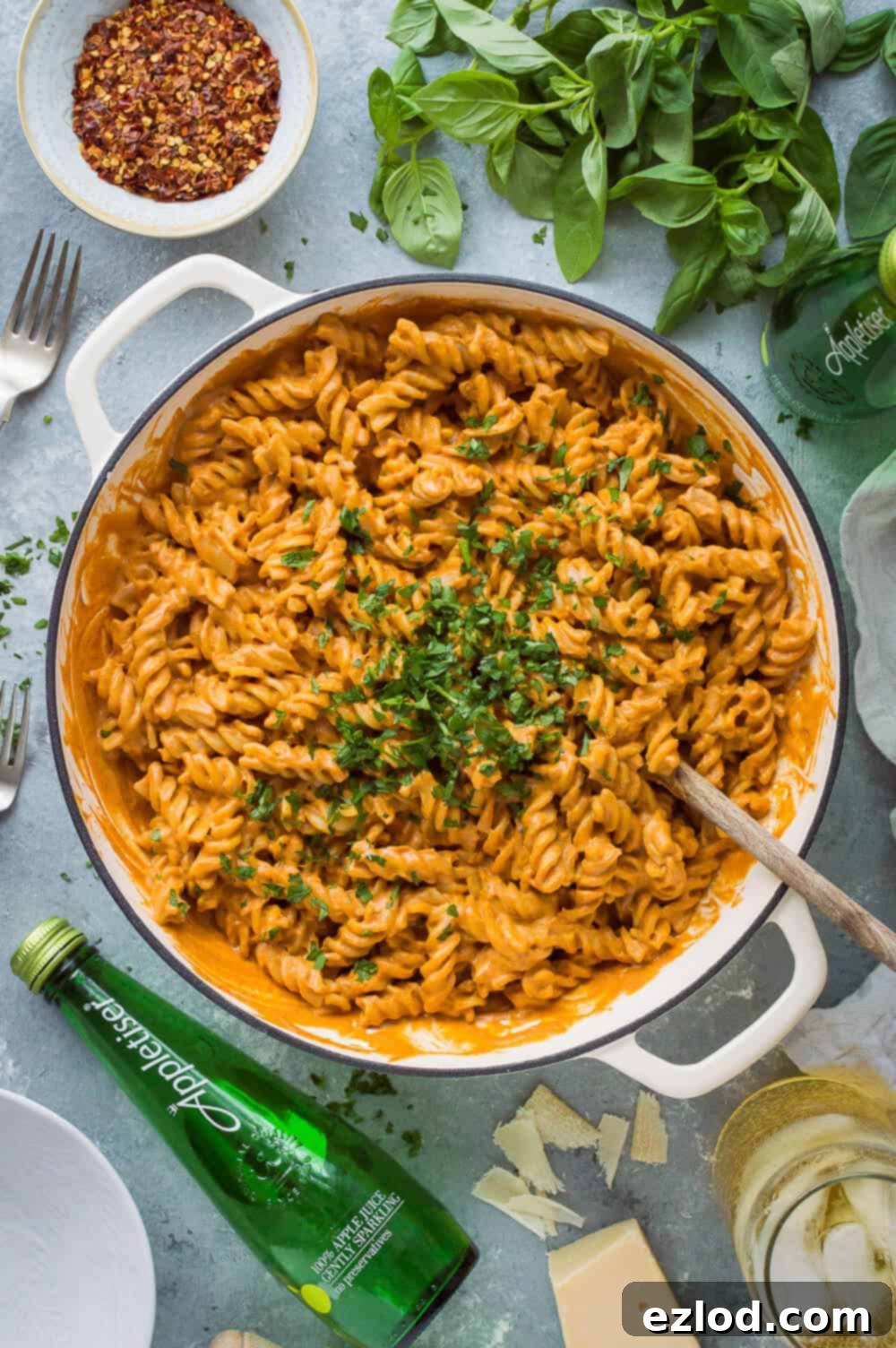 Flatlay of a pan of vegan creamy roasted red pepper tomato pasta on a grey background with herbs, cutlery and Appletiser.