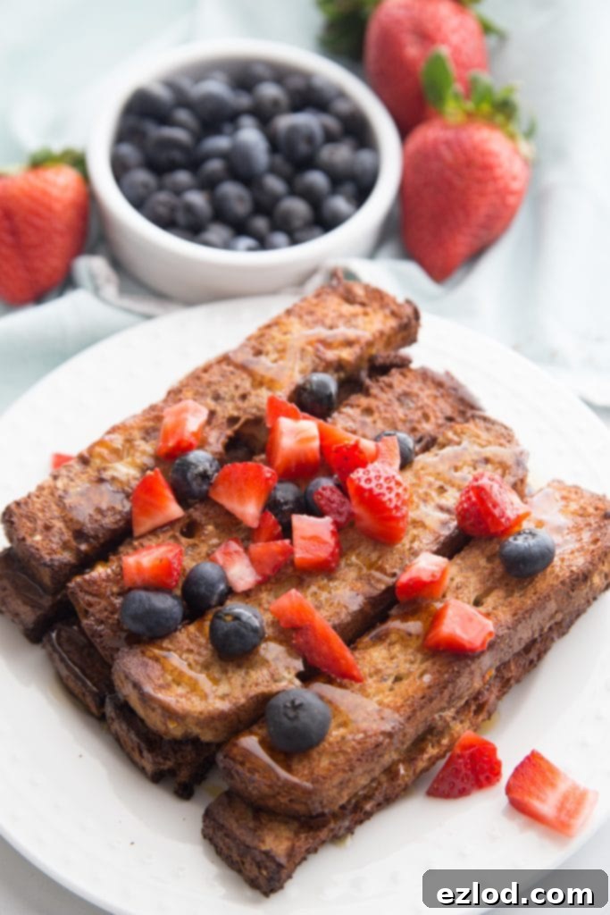 A stack of golden-brown Healthy Air Fryer French Toast Sticks topped with fresh strawberries and blueberries on a white plate, ready to be served for breakfast.