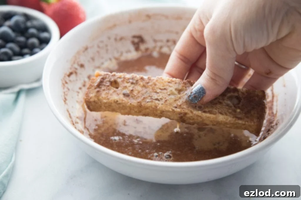 A French toast stick, perfectly sliced from a hearty piece of bread, is being gently dipped into a rich, golden egg mixture in a shallow bowl, ensuring full saturation before cooking.