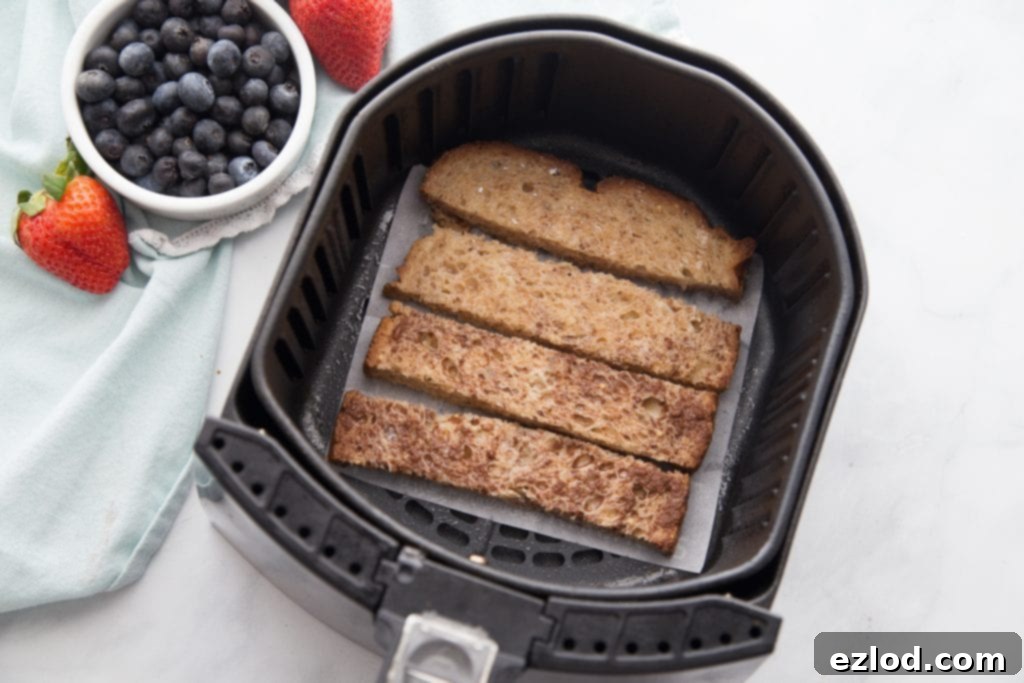 A close-up view inside an air fryer basket, revealing a single layer of perfectly golden-brown French toast sticks, lightly sprayed with cooking oil, ready for air frying.