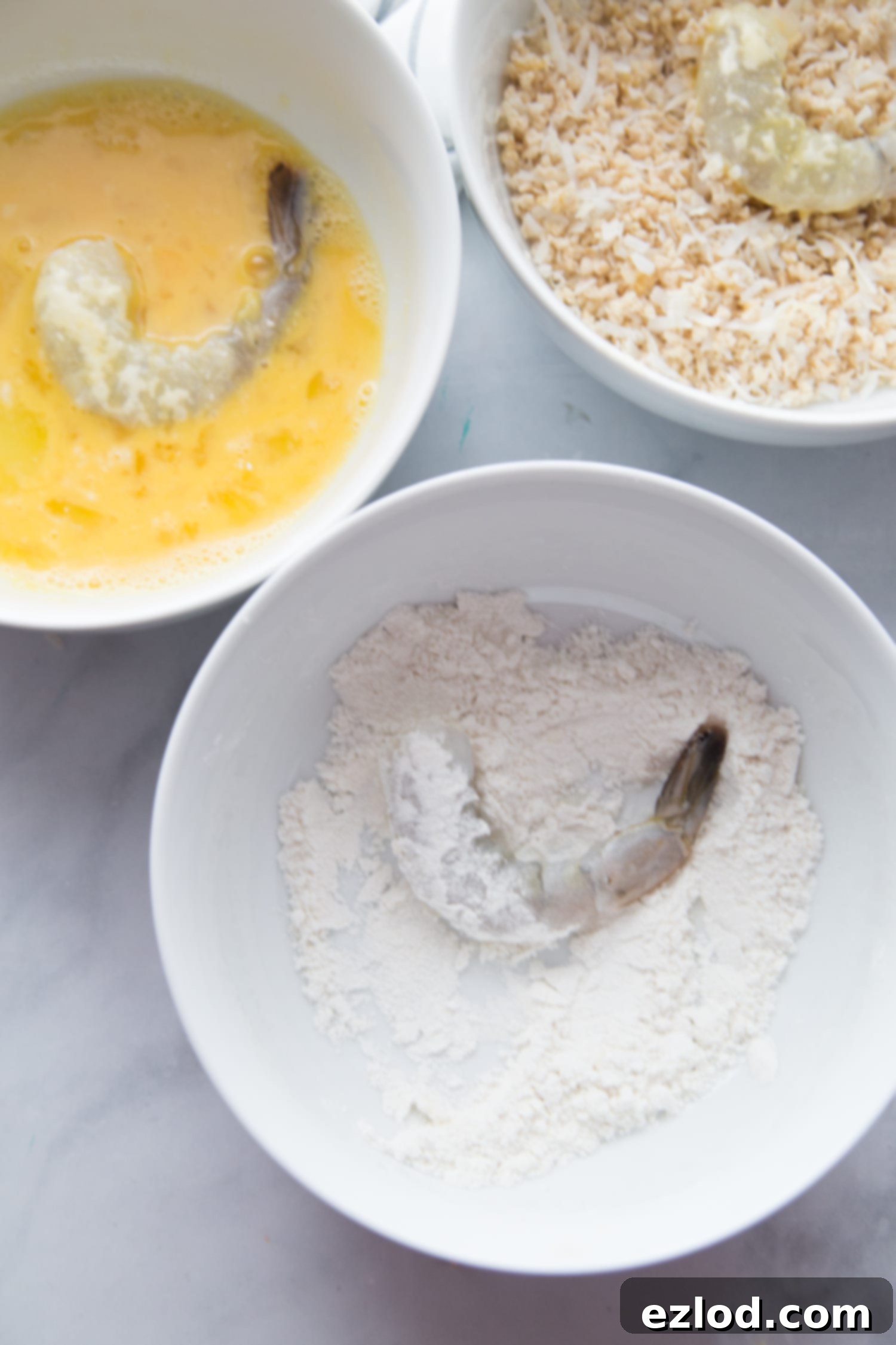 Three white bowls on a marble backdrop - one has egg with a shrimp, one has gluten free flour with shrimp, and one has panko breadcrumb with shrimp. 