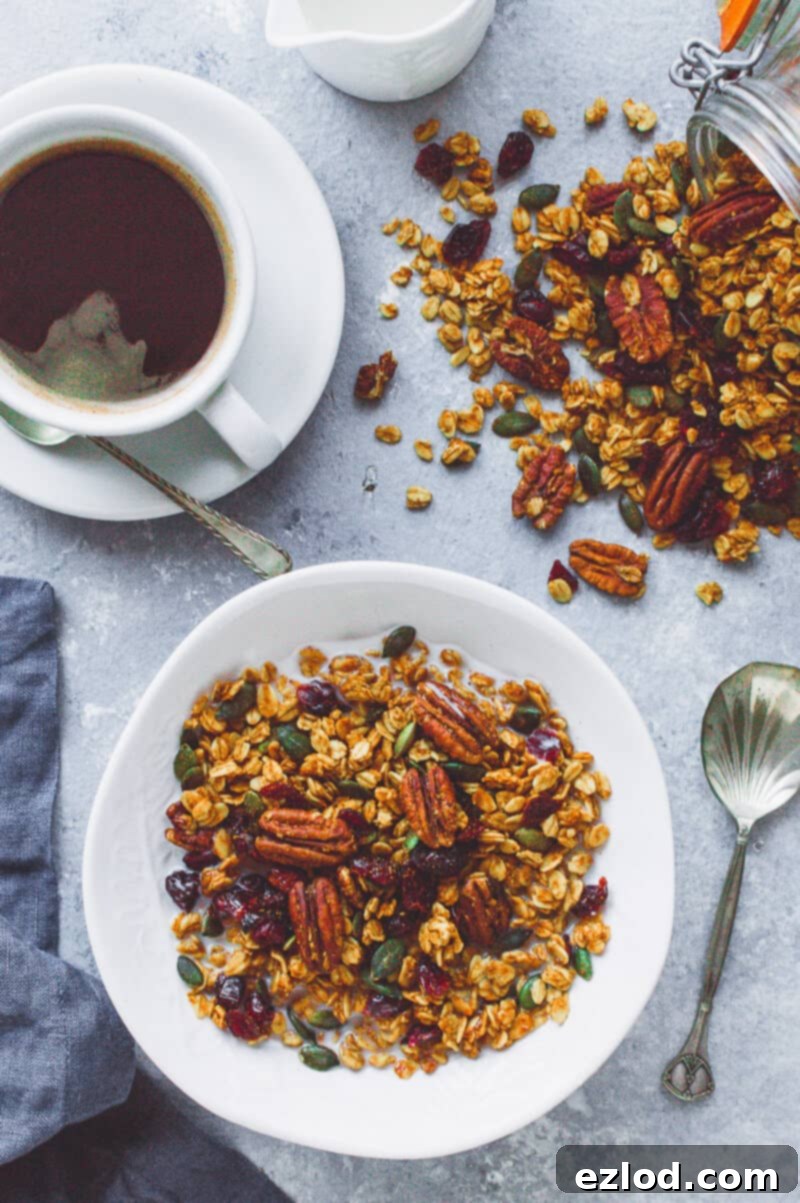 A bowl of vegan pumpkin spice granola with coffee, a jug of milk and a jar of spilled granola.