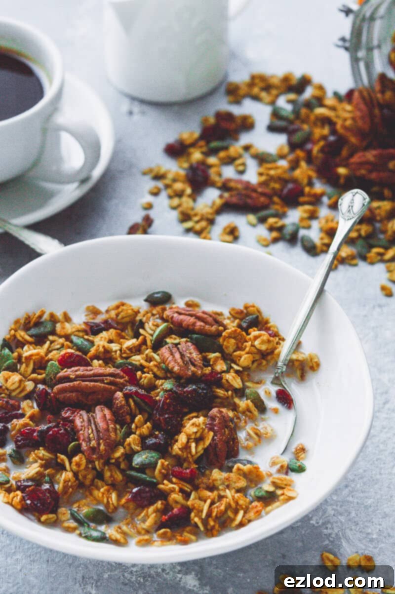 Close up of a bowl of vegan pumpkin spice granola with milk, with coffee and spilled granola in the background.