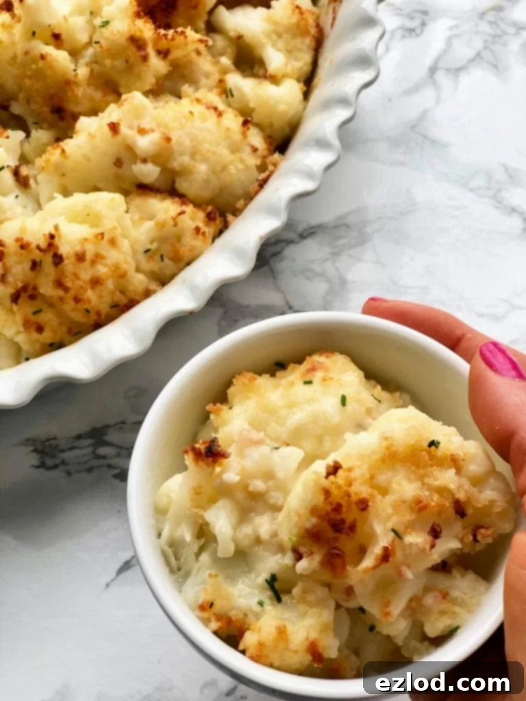 A woman's hand is holding a small bowl of Cauliflower Au Gratin next to a larger casserole dish of the same recipe. 