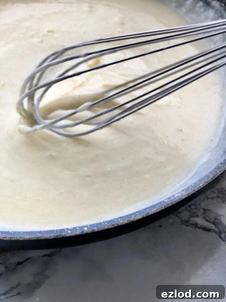 A white creamy cheese sauce is being whisked in a skillet using a wire whisk 