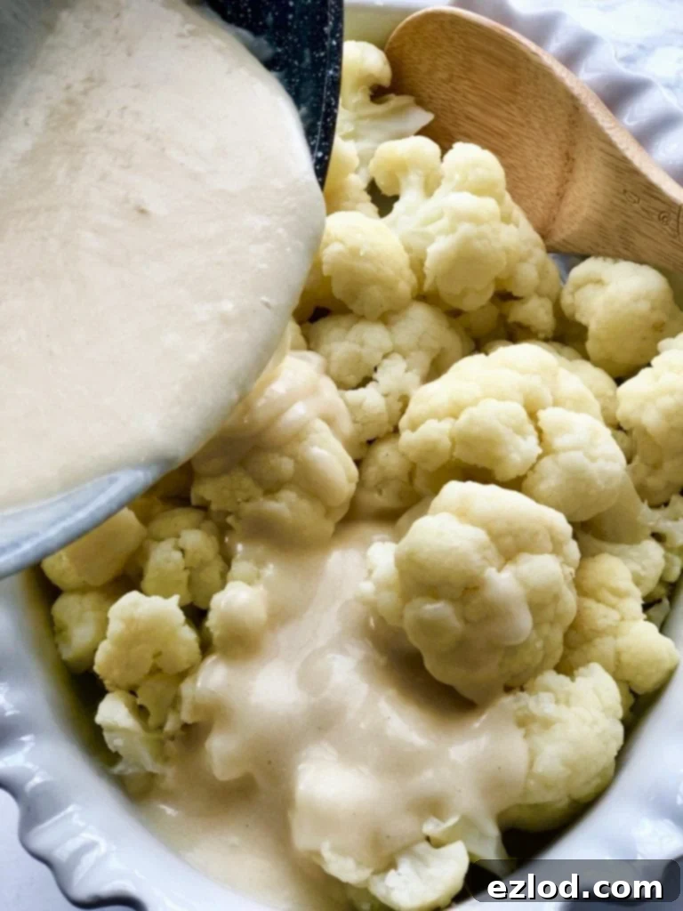 A white creamy cheese sauce is being poured over cauliflower florets in a white oval casserole dish