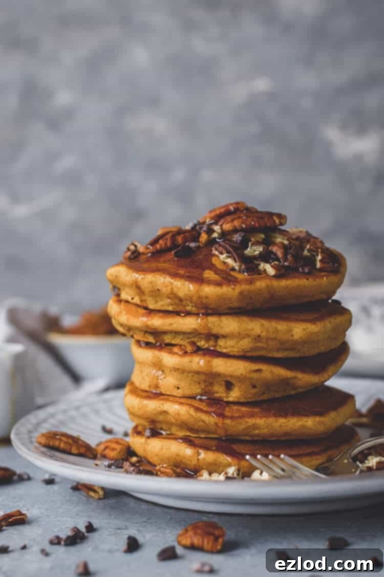 A stack of golden brown, fluffy vegan pumpkin pancakes drizzled with maple syrup, garnished with toasted pecans and cacao nibs, on a white plate, ready for a cozy autumn breakfast. #vegan #pumpkin #pumpkinspice #pancakes #brunch