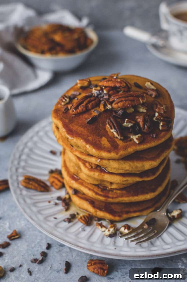 A beautifully composed shot of a stack of fluffy vegan pumpkin pancakes on a white plate, generously topped with golden maple syrup, crunchy pecans, and dark cacao nibs, highlighting their delicious appeal.