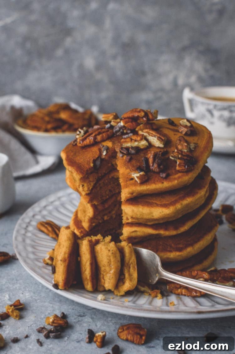 A close-up of a stack of fluffy vegan pumpkin pancakes, one forkful taken out to reveal the soft interior. Topped with maple syrup, toasted pecans, and cacao nibs, capturing the irresistible appeal of a cozy autumn breakfast.