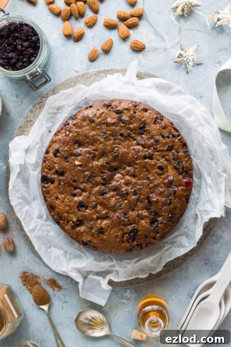 An overhead shot of a prepared vegan Christmas fruitcake on white parchment paper, surrounded by various essential ingredients like dried fruits, fragrant spices, and crunchy almonds, ready for the baking process.