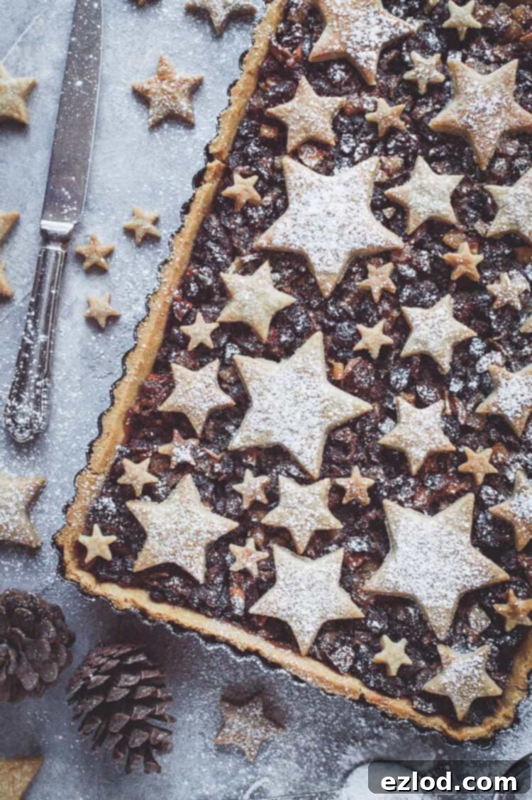 The fully baked vegan shortbread star topped mince pie tart elegantly displayed on a grey background, surrounded by various festive Christmas decorations. This beautiful dessert is ready to be served as a stunning holiday centrepiece.