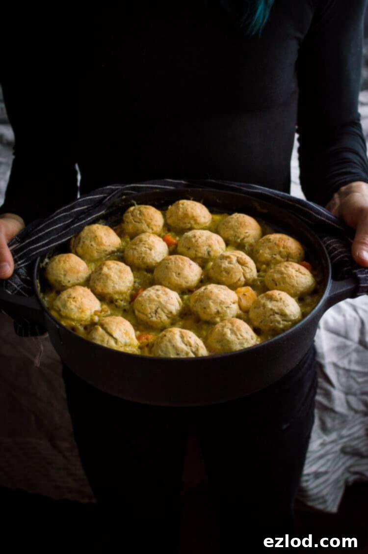 A person in black attire holding a black cast iron pot filled with a finished vegan vegetable and pearl barley stew, showcasing the herby dumplings on top, emphasizing the rustic and homemade feel of the dish.