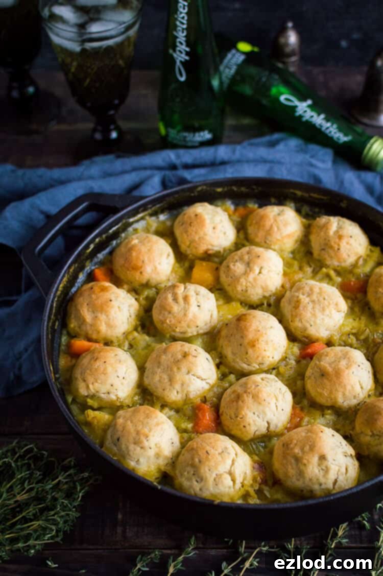 An overhead shot showcasing a rustic cast iron pot brimming with a rich vegan vegetable and pearl barley stew, adorned with a scattering of golden herby dumplings. A bottle of Appletiser stands elegantly in the background, suggesting a perfect meal pairing.