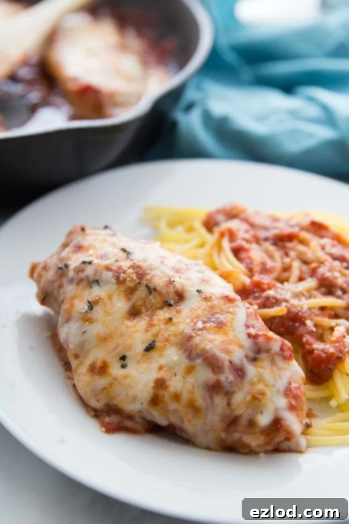 Chicken breast topped with tomato sauce and melted cheese next to spaghetti and tomato sauce on a white plate, with the rest of the Italian chicken skillet in the background
