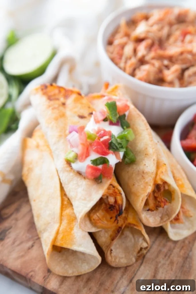 Five crispy chicken taquitos with fresh toppings stacked in a pyramid, with extra salsa lime chicken in a white bowl in the background