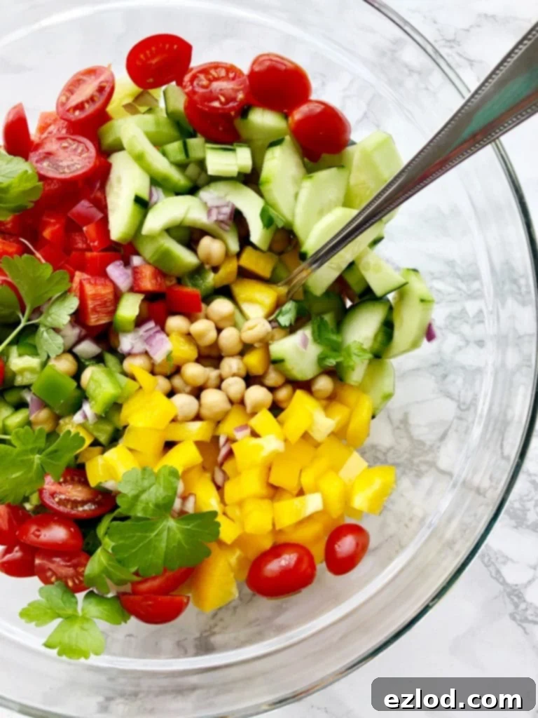 Lots of colorful bell peppers, grape tomatoes, and cucumbers in a bowl with drained chickpeas, ready to be mixed into a vibrant Greek Chickpea Salad.
