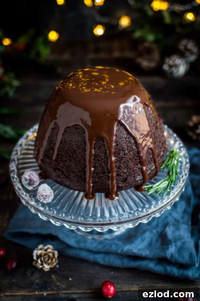 A beautifully presented slice of vegan steamed chocolate pudding, generously drizzled with rich chocolate sauce, resting on a glass plate atop a rustic wooden background.