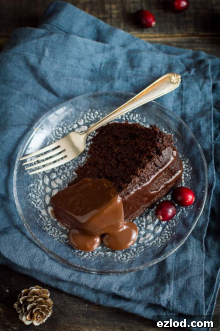 An inviting slice of vegan steamed chocolate pudding, generously topped with a decadent chocolate sauce, artfully arranged on a glass plate against a vibrant blue cloth.