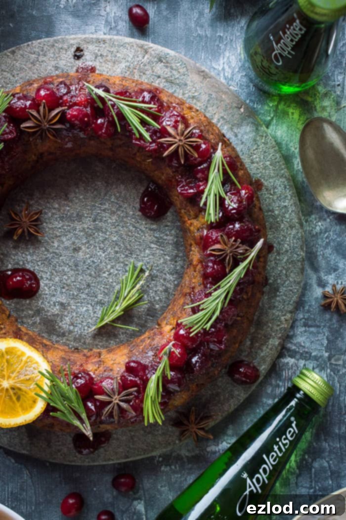 Close-up of a festive vegan nut roast wreath adorned with vibrant cranberry sauce, fresh rosemary, and whole spices, with an Appletiser bottle in the background, signifying a perfect pairing.
