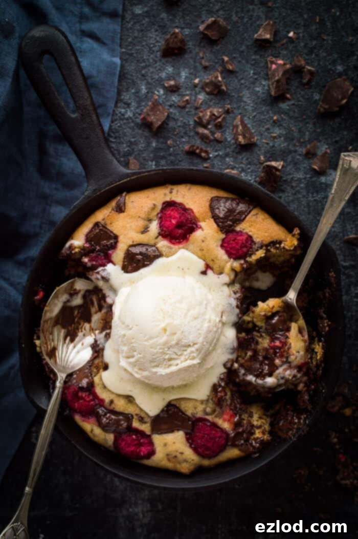 Partially eaten mini vegan chocolate raspberry skillet cookie for two topped with vanilla ice cream with two spoons on a black background.