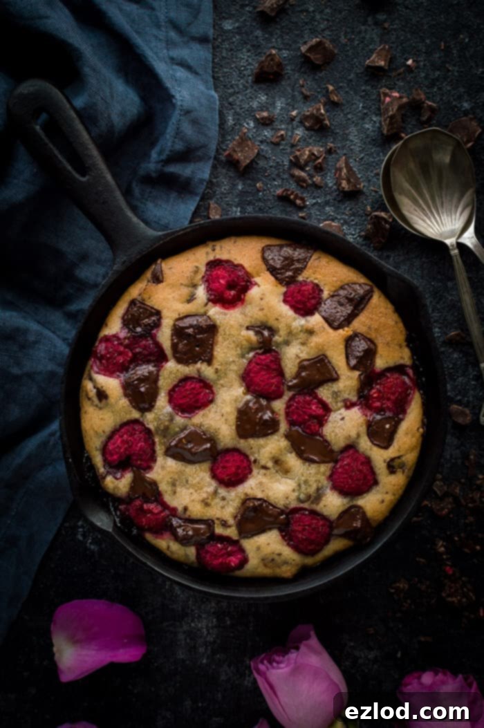 Chocolate raspberry skillet cookie for two on a black background with two spoons and a grey cloth.