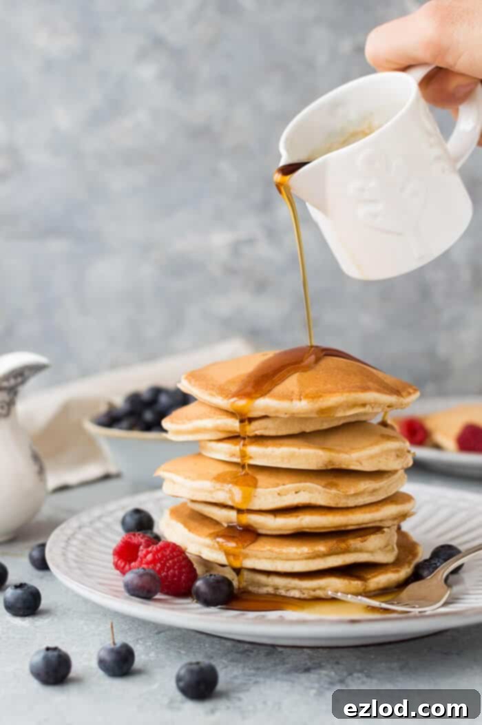 Maple syrup being generously poured over a tall stack of golden brown fluffy vegan vanilla pancakes on a plate, ready to be enjoyed.