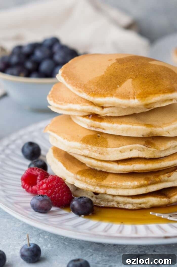 A close-up of a perfectly styled stack of fluffy vegan vanilla pancakes, topped with maple syrup and an assortment of fresh berries, elegantly presented on a white plate.