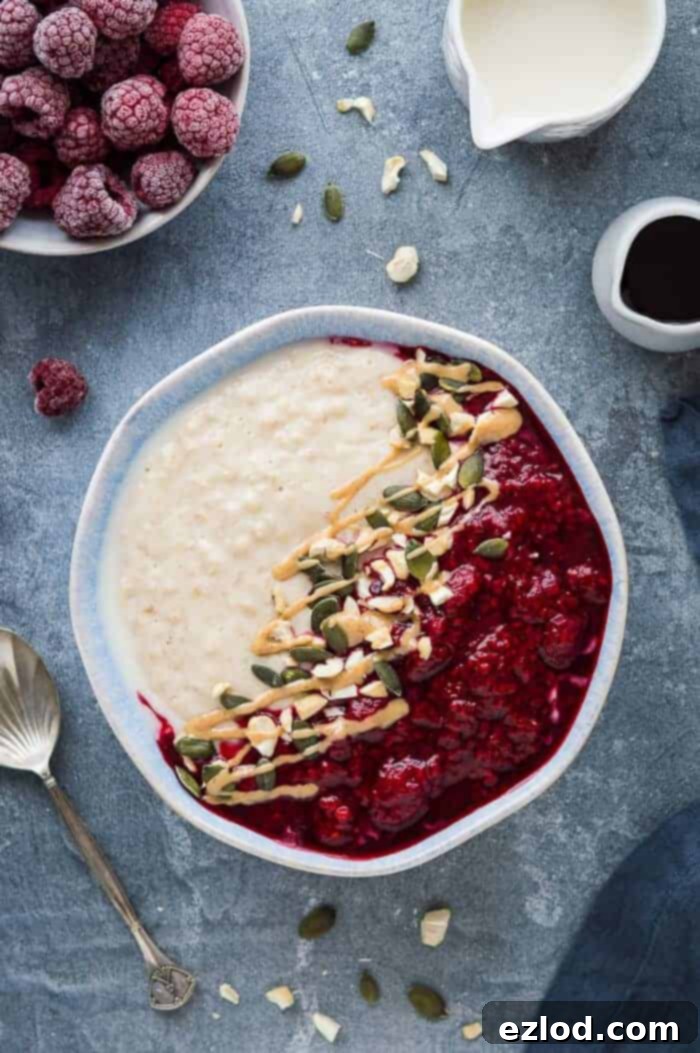 Vegan peanut butter porridge topped with tangy raspberry compote, seeds and peanut butter in a blue bowl on a grey background with frozen raspberries, a jug of milk, maple syrup and a spoon.