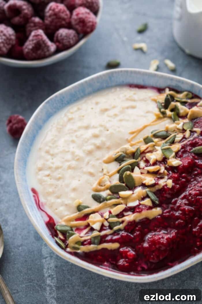 Side shot of a bowl of vegan peanut butter and raspberry porridge with a bowl of frozen raspberries and a jug of milk in the background.