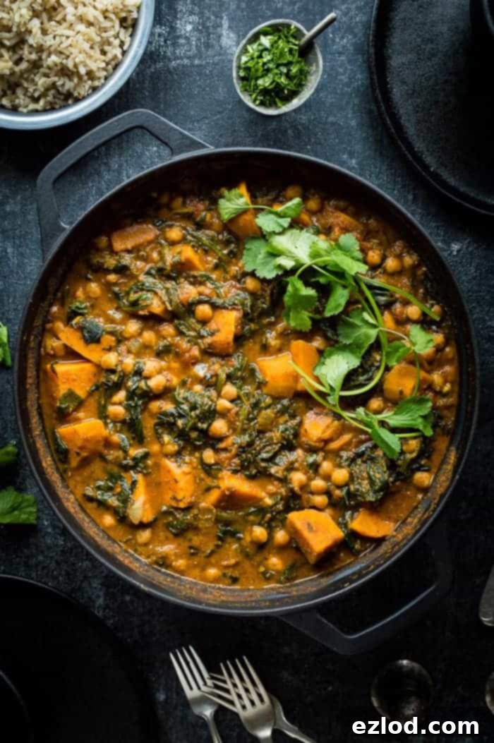 A large black cast iron pan filled with a steaming, vibrant vegan chickpea, sweet potato, and spinach curry, ready to be served.