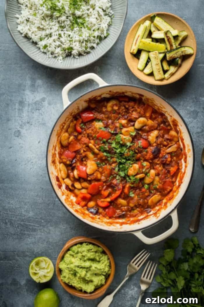 A vibrant pan of vegan vegetable, lentil, and butter bean chilli, accompanied by a bowl of fresh coriander rice, tender roasted courgettes, and a dollop of creamy guacamole, all set against a modern grey background.